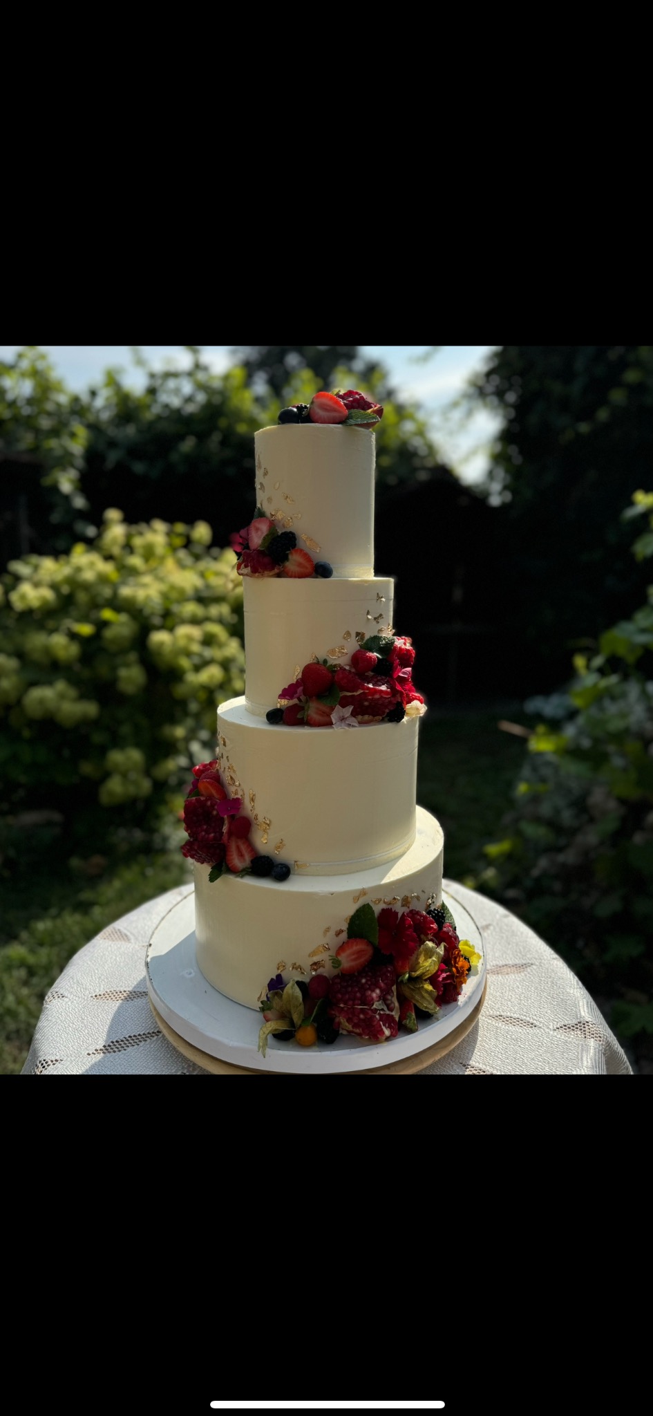 Elegancki, czteropiętrowy tort weselny z białym kremem, zdobiony świeżymi owocami (truskawki, borówki, maliny, granat) i płatkami złota na tle zieleni.