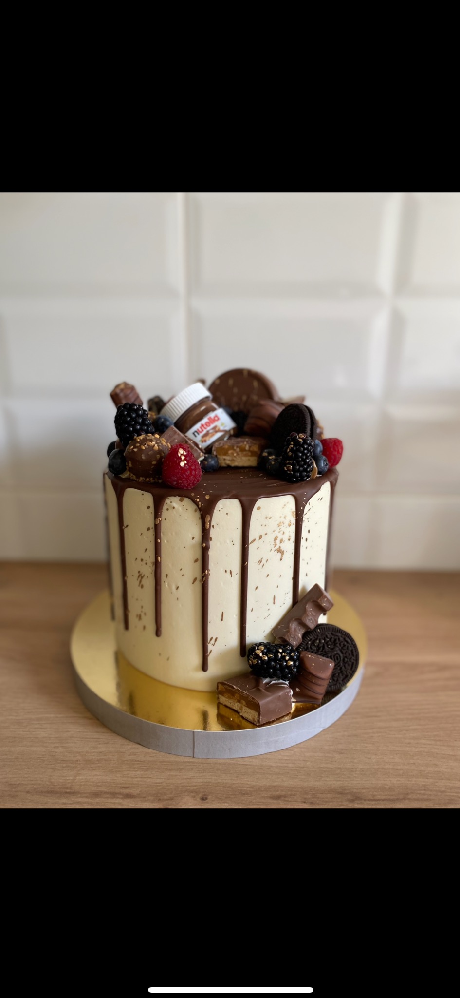 Tort z polewą czekoladową, ozdobiony owocami, batonami i mini słoikiem Nutelli, na złotym podkładzie, na drewnianym blacie, z białymi płytkami w tle.