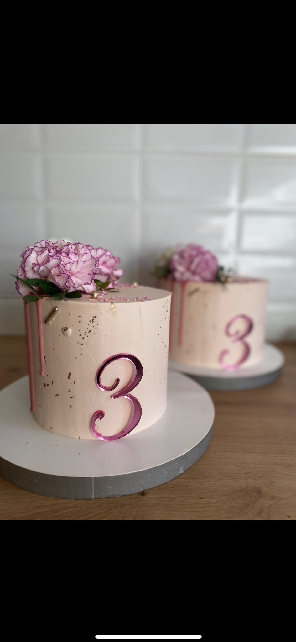 Dwa torty urodzinowe w Niegowonicach z kremowym lukrem, kwiatami i cyfrą 3. Jeden tort jest blisko, drugi w tle, oba na drewnianym blacie.