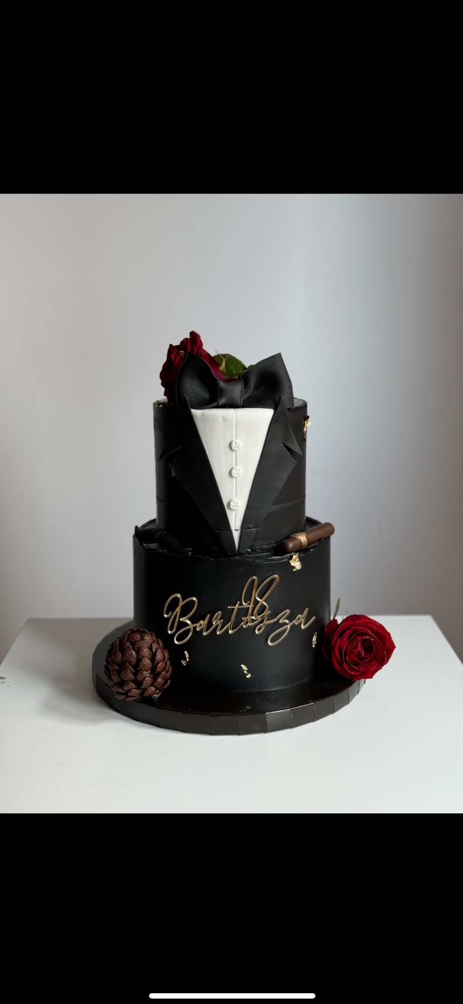 Dwupoziomowy tort w stylu smokingowym, zdobiony czerwoną różą i złotymi płatkami, z napisem 'Bartosza'. Elegancka kompozycja na jasnym tle.