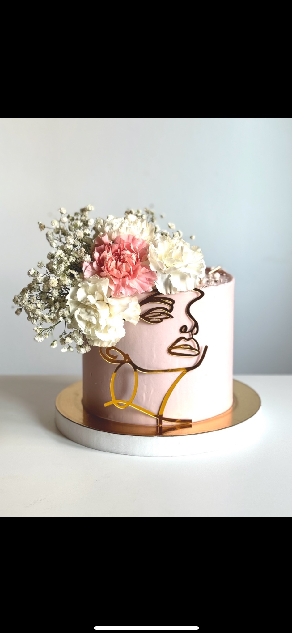 Różowy tort zdobiony kwiatami i złotym konturem twarzy, na złotym podeście. Elegancki i artystyczny tort na specjalną okazję.