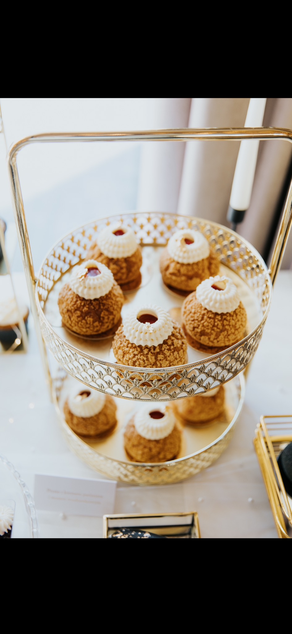 Elegancka, dwupoziomowa etażerka ze złotego metalu prezentuje kremowe ptysie z dekoracją z bitej śmietany i dżemu na jasnym tle. Delikatne oświetlenie.