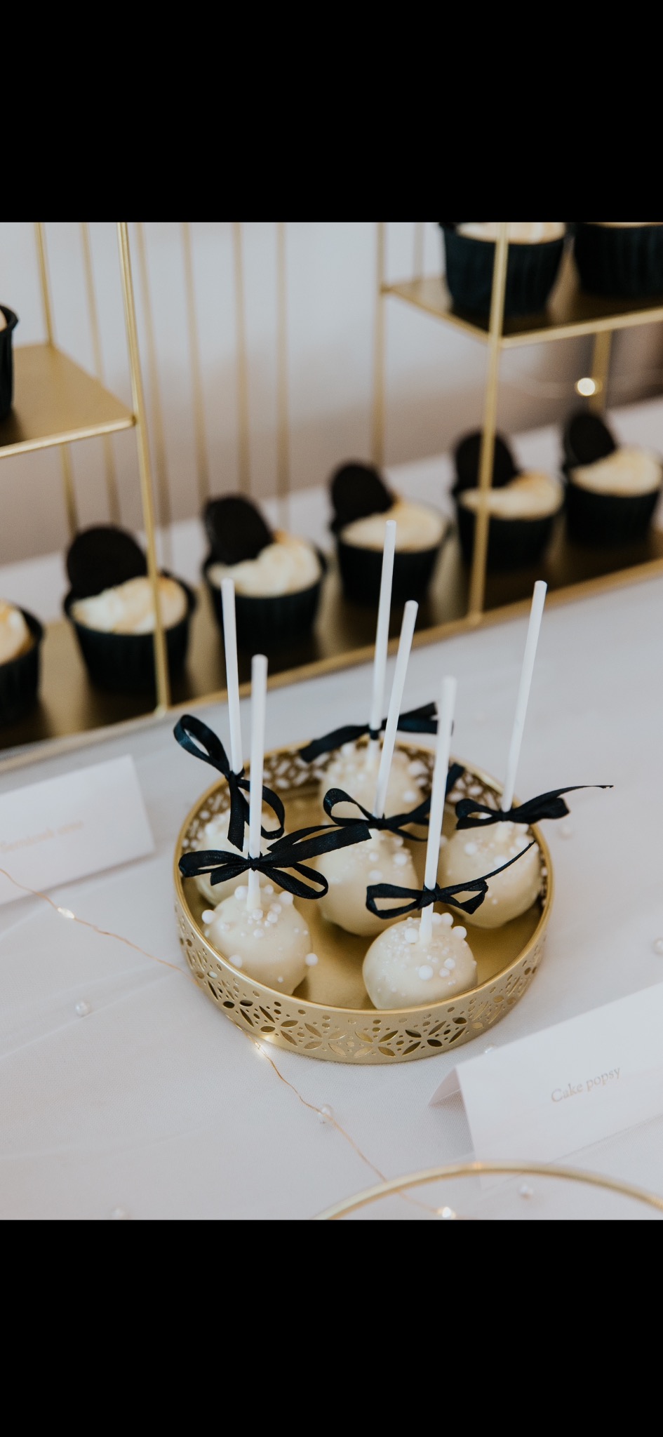 Eleganckie cake pops z białej czekolady, zdobione perełkami i czarnymi kokardkami, na złotym talerzu. W tle muffiny i dekoracje.
