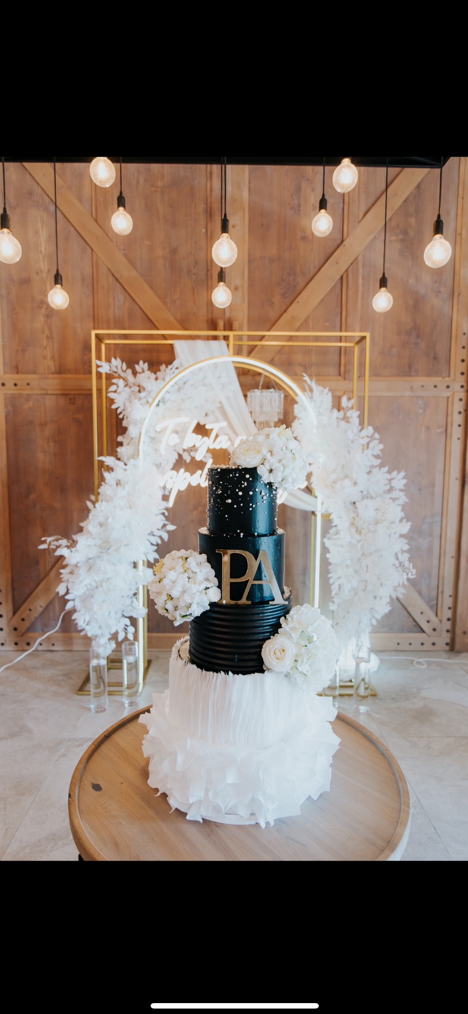 Elegancki, czteropiętrowy tort weselny w czarno-białej kolorystyce, udekorowany kwiatami i złotymi inicjałami 'PA', na drewnianym stole, z dekoracyjnym tłem.