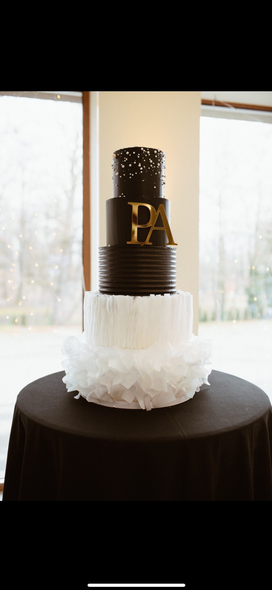 Elegancki, czteropiętrowy tort weselny w odcieniach czerni i bieli, udekorowany złotymi inicjałami 'PA' i umieszczony na czarnym obrusie.
