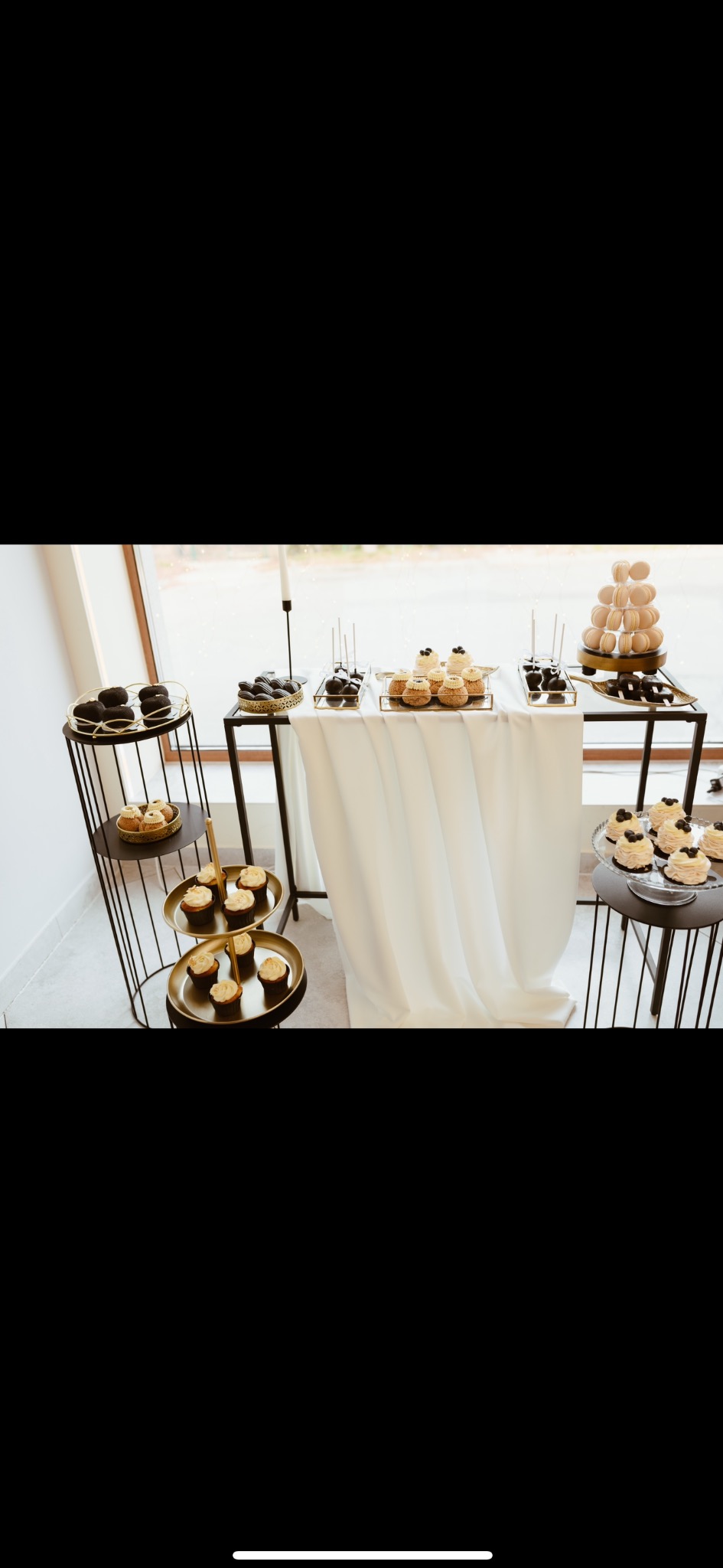 Stół z białym obrusem i czarne, metalowe etażerki prezentują różnorodne desery: babeczki, cake popsy, makaroniki i inne słodkości. Elegancki wystrój.