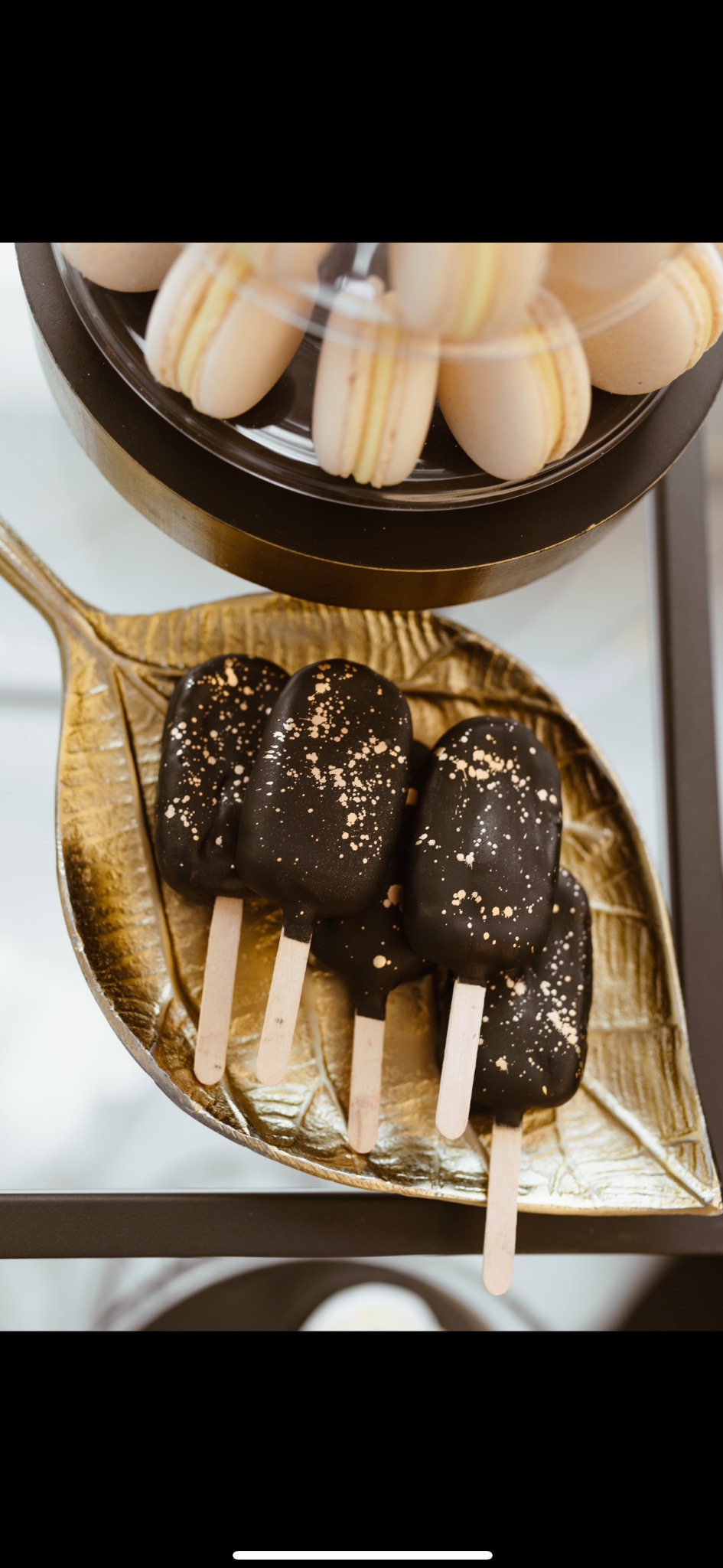 Czekoladowe cake pops z posypką na złotej paterze w kształcie liścia, obok makaroniki w przezroczystym pudełku. Eleganckie słodkości.
