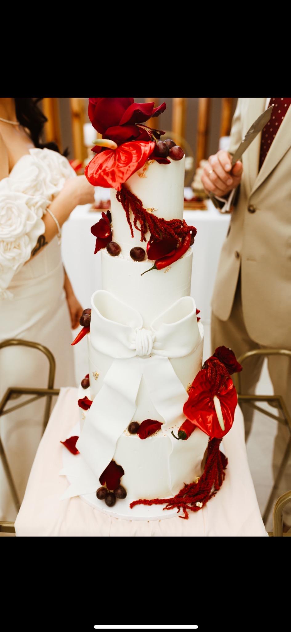 Elegancki, trzypiętrowy tort weselny z białym lukrem, zdobiony czerwonymi kwiatami anturium, winogronami i dużym białym lukrowym kokardą. Nóż w dłoni osoby w garniturze.