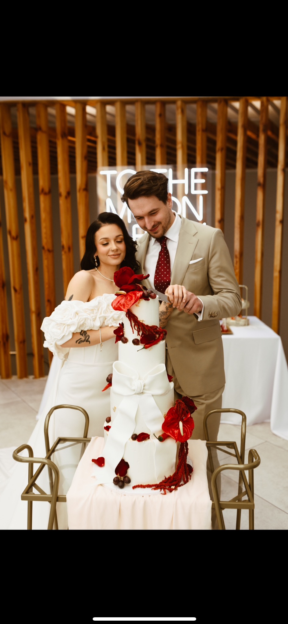 Elegancki, trzypiętrowy tort weselny z białym lukrem i czerwonymi kwiatami, krojony przez parę młodą na tle drewnianej ściany z neonowym napisem.