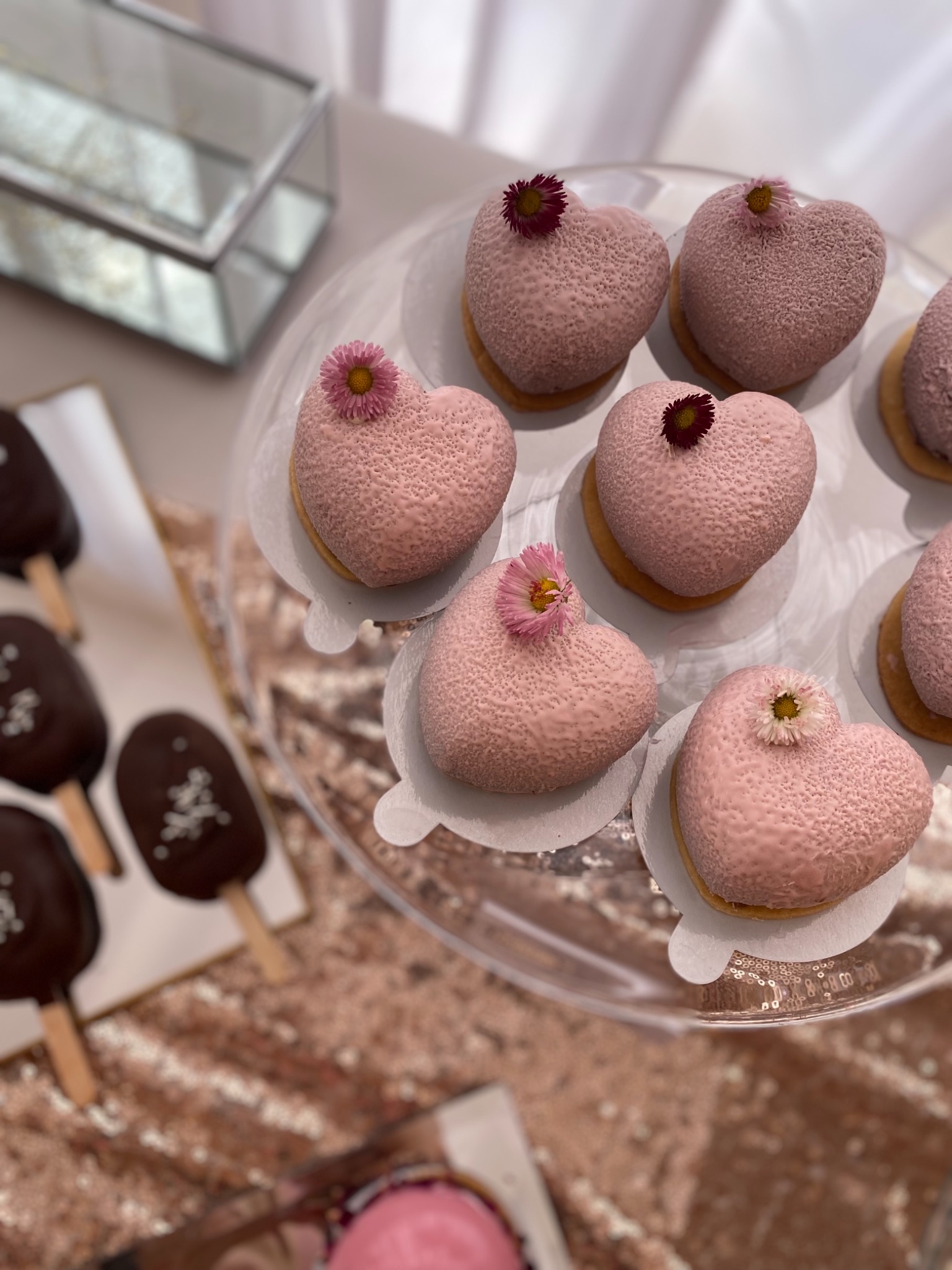 Różowe desery w kształcie serca z jadalnymi kwiatami na przezroczystym talerzu, obok czekoladowych lodów na patyku i dekoracyjnej szkatułki.