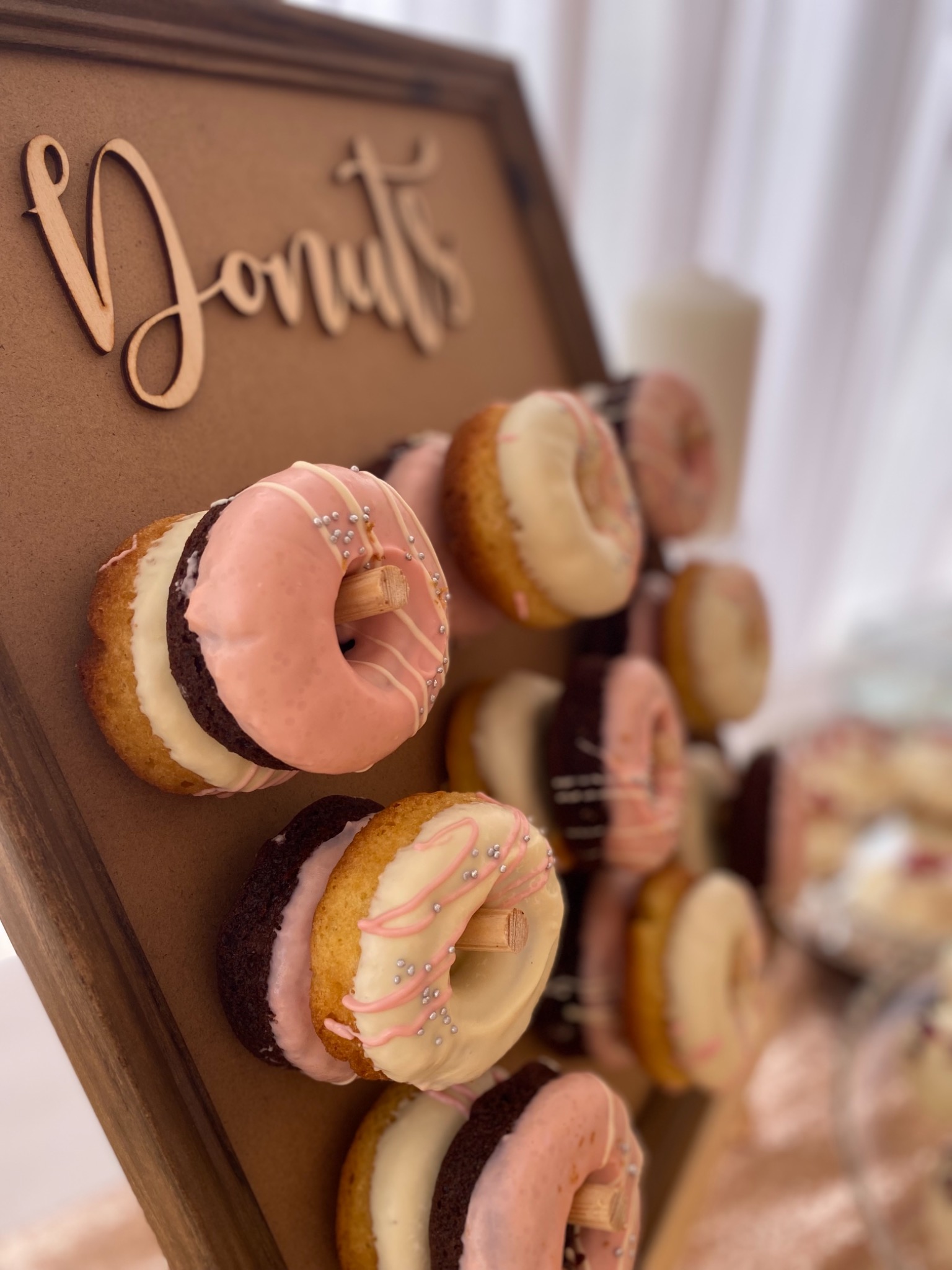 Drewniany stojak z napisem 'Donuts' prezentujący dekorowane pączki w różnych smakach (czekoladowy, waniliowy, różowy) na drewnianych kołkach, ustawiony pod kątem.