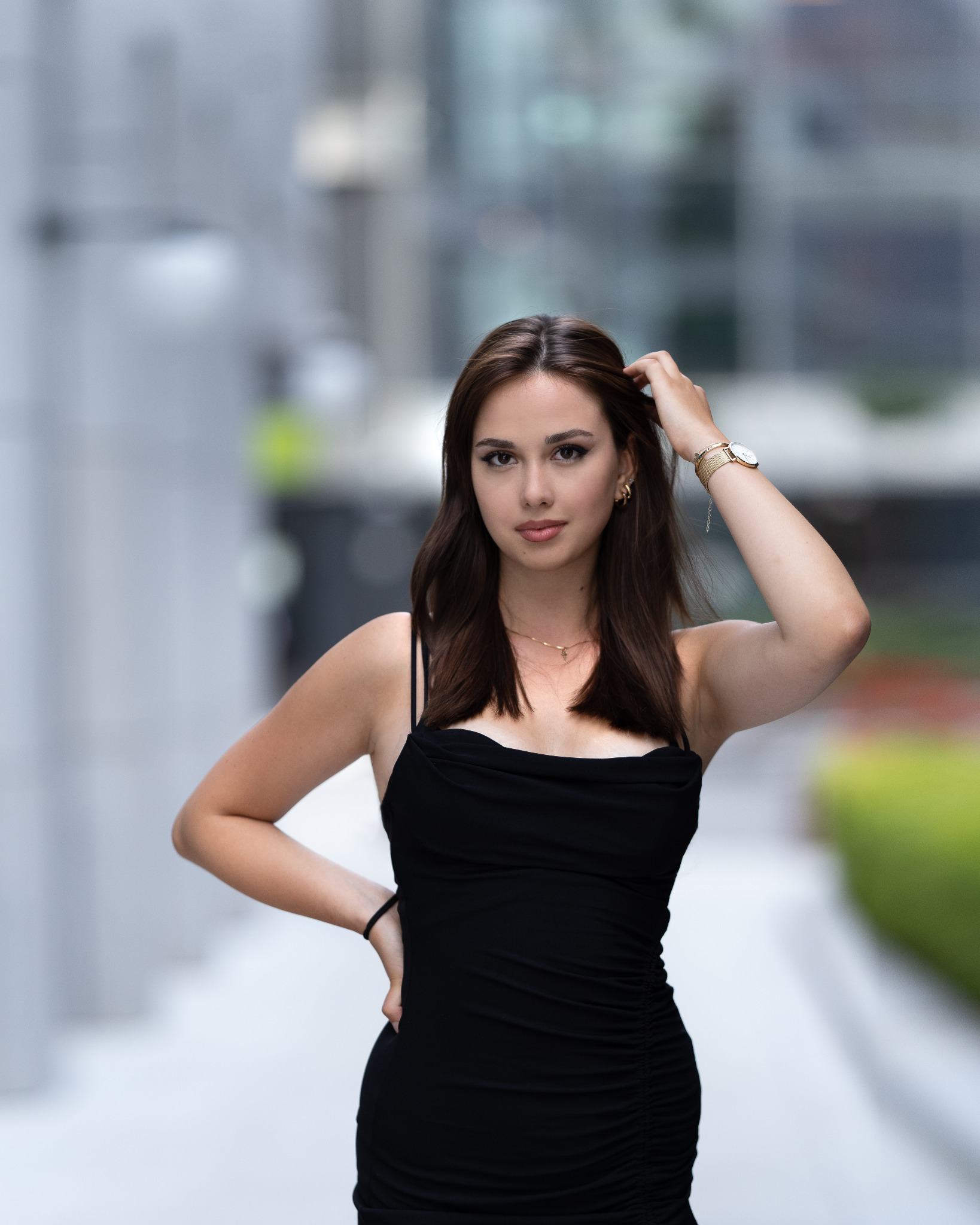 Portret młodej kobiety w czarnej sukience, pozującej na tle nowoczesnej architektury miejskiej, z ręką we włosach i złotym zegarkiem na nadgarstku.