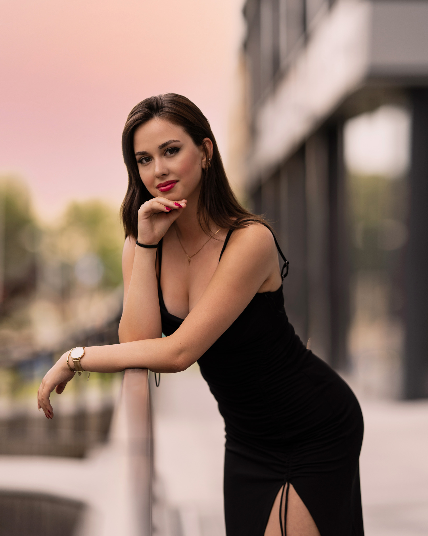 Portret kobiety w czarnej sukience, opierającej się o barierkę na tle miejskiego krajobrazu z pastelowym niebem, z widocznym zegarkiem na ręce i delikatną biżuterią