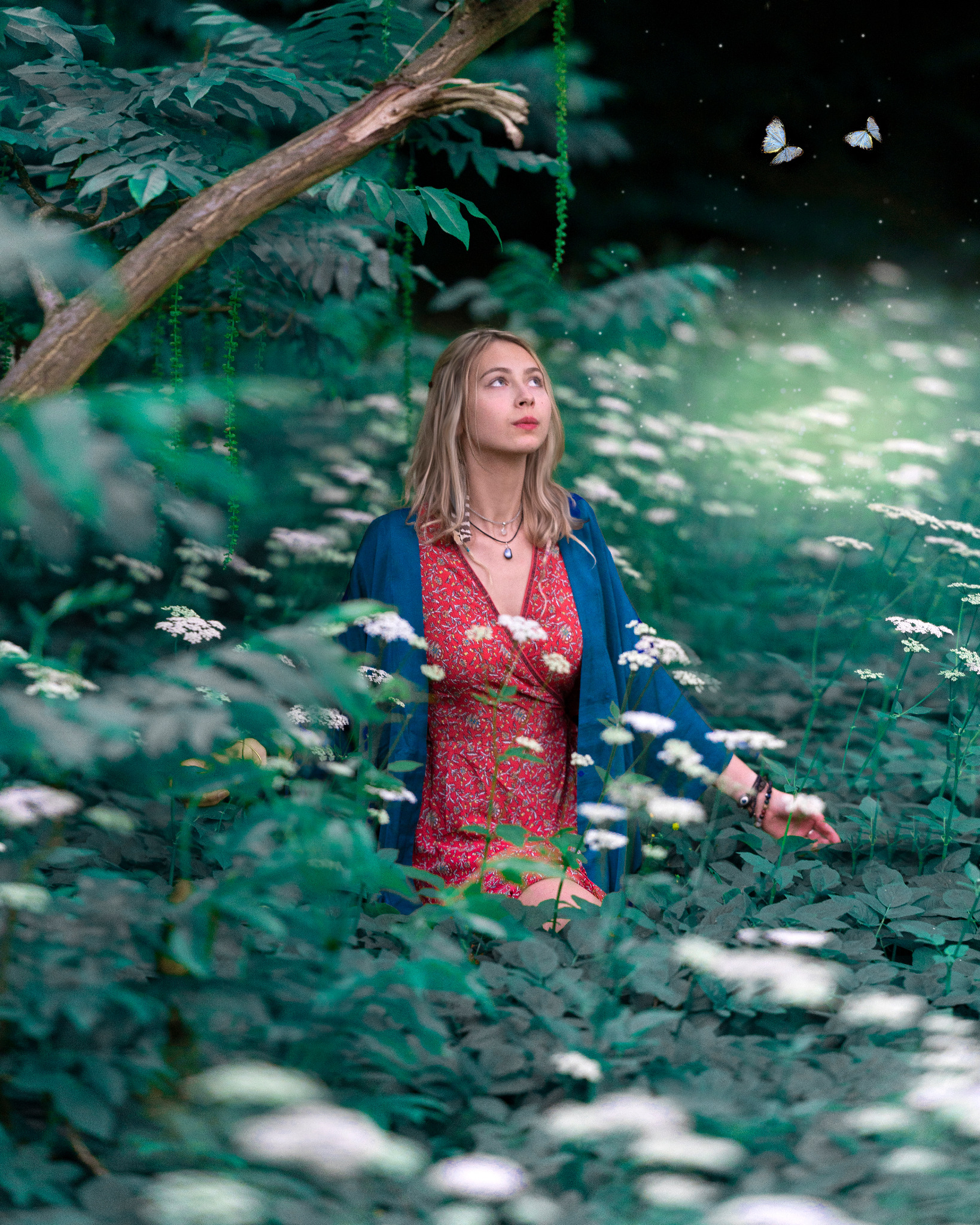 Kobieta w kwiecistej sukience i niebieskim kardiganie stoi pośród białych kwiatów i zieleni, spoglądając w górę na dwa motyle na tle ciemnego nieba z gwiazdami.