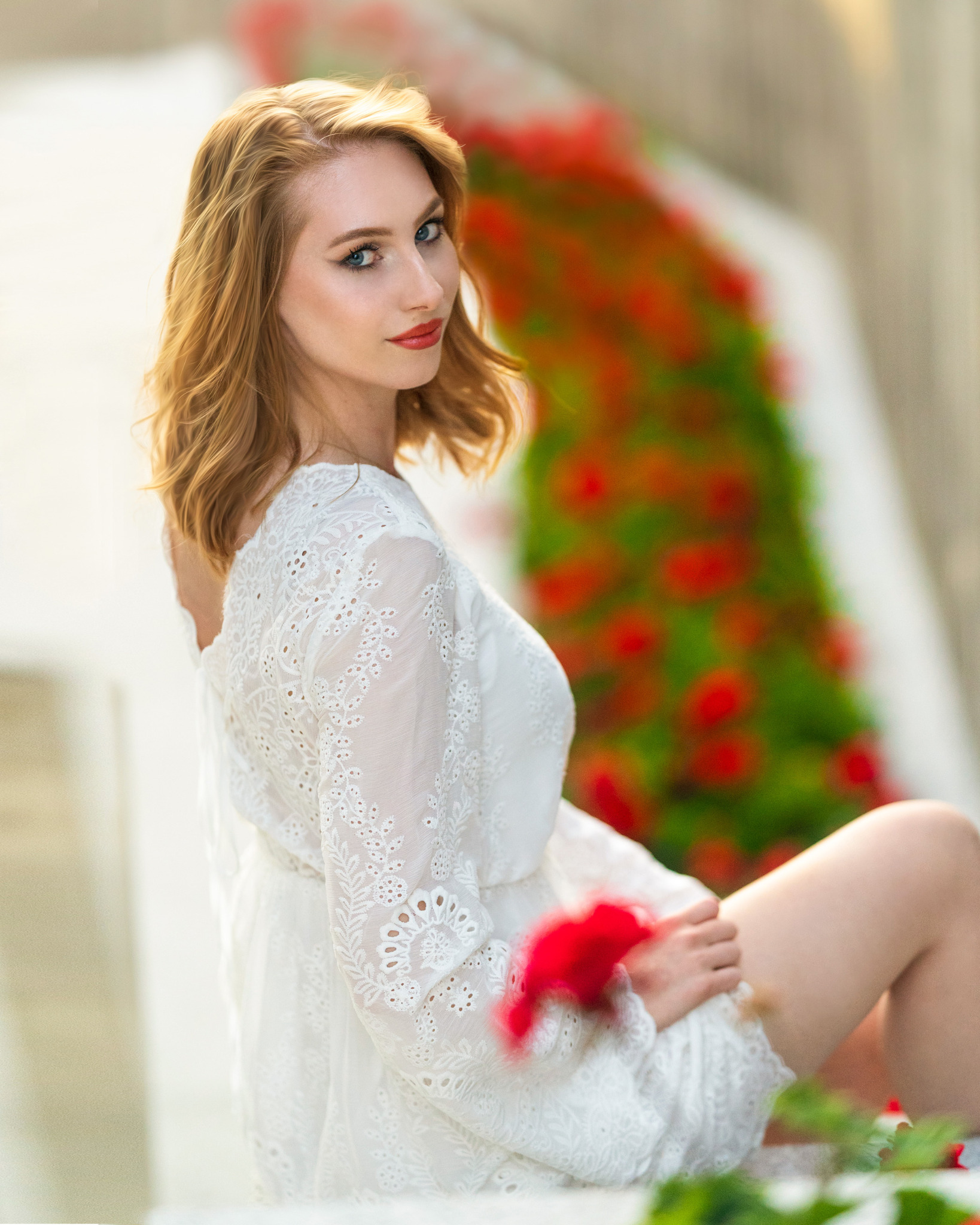 Portret kobiety w białej, ażurowej sukience, siedzącej na tle ściany z czerwonymi kwiatami.