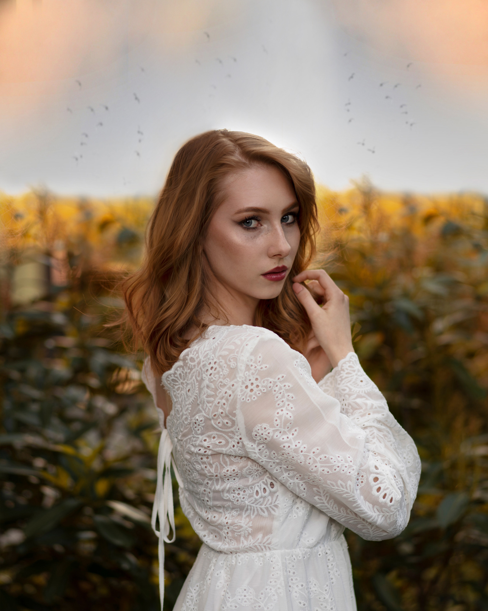 Portret kobiety o rudych włosach w białej, ażurowej sukience, pozującej na tle żółto-zielonego pola, z widocznymi ptakami na niebie.