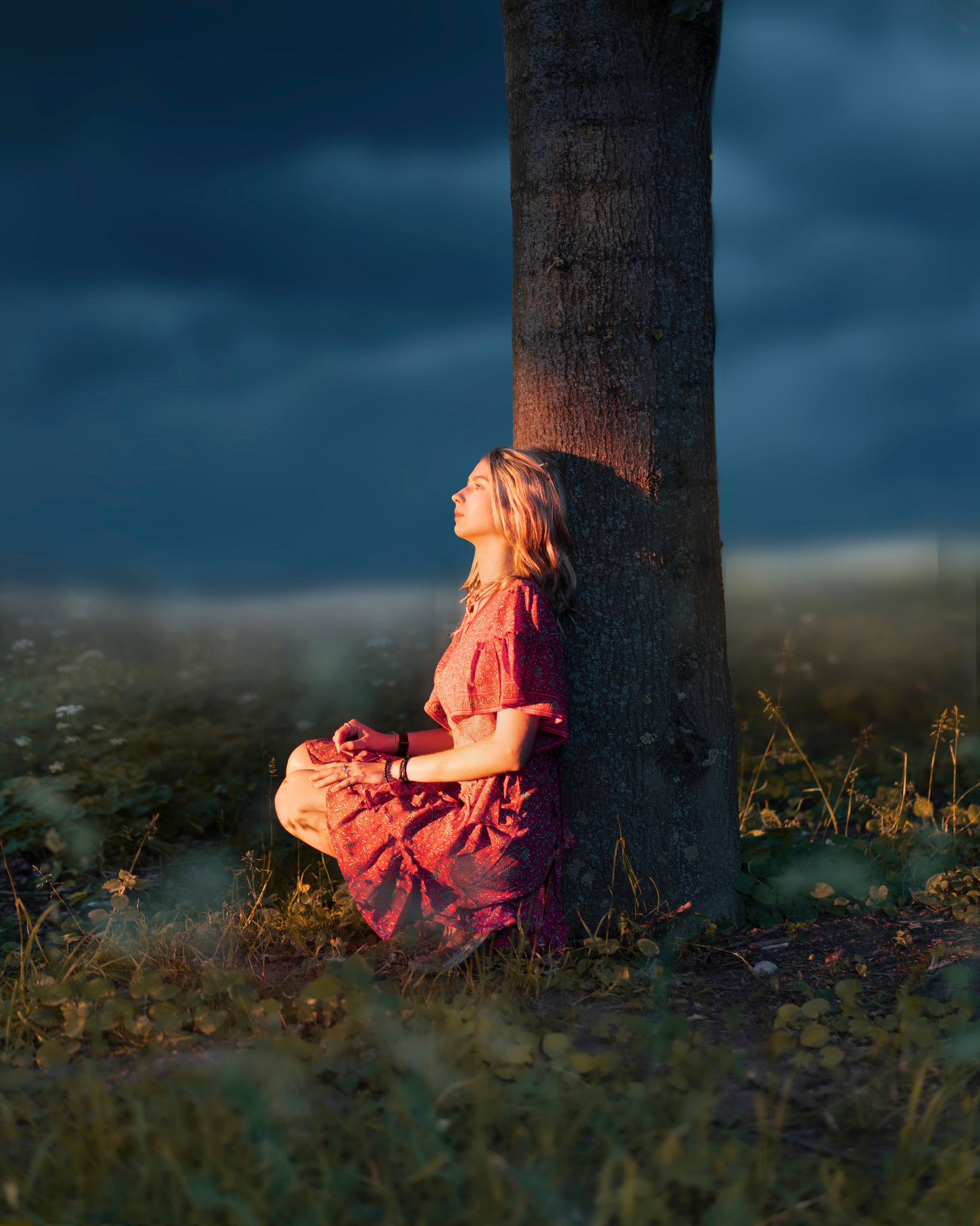 Kobieta w kwiecistej sukience kuca oparta o pień drzewa, spoglądając w górę, oświetlona ciepłym, zachodzącym słońcem na tle ciemnego, rozmytego nieba.