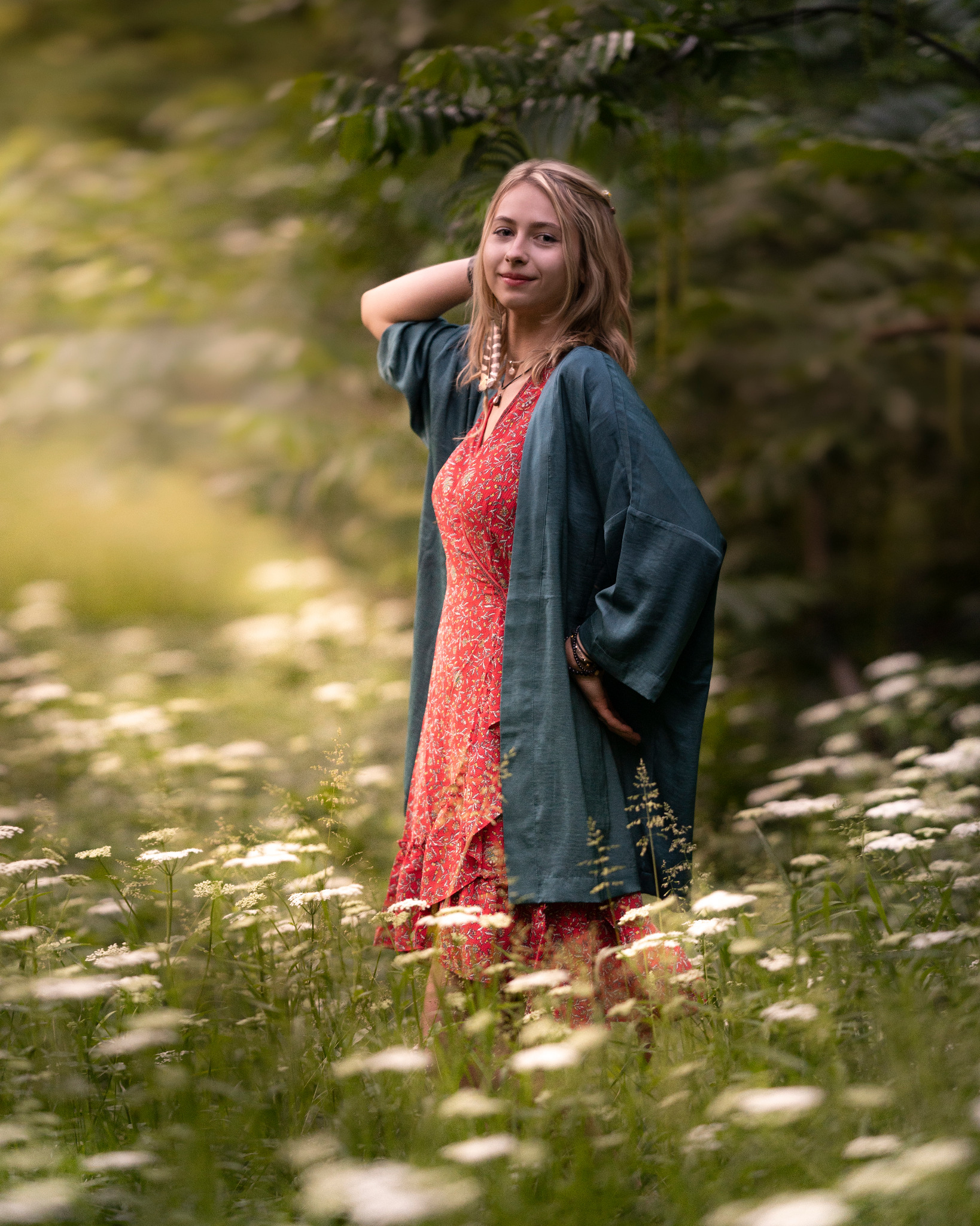 Młoda kobieta w czerwonej sukience i zielonej narzutce pozuje w kwitnącej łące, dłoń uniesiona za głową, delikatny uśmiech, naturalne światło.