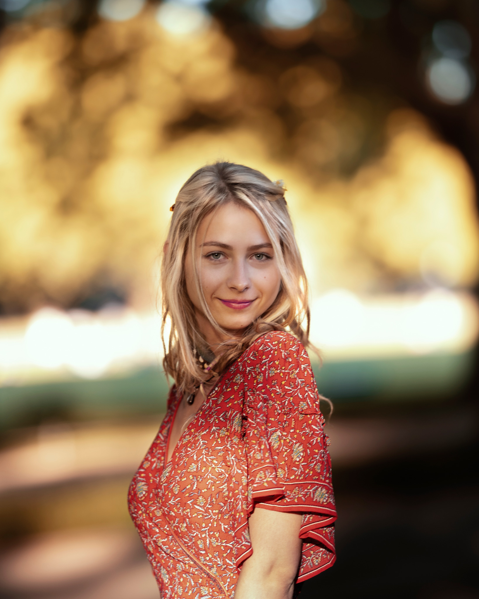 Portret uśmiechniętej kobiety w czerwonej kwiecistej sukience na tle rozmytej zieleni i złotego światła.