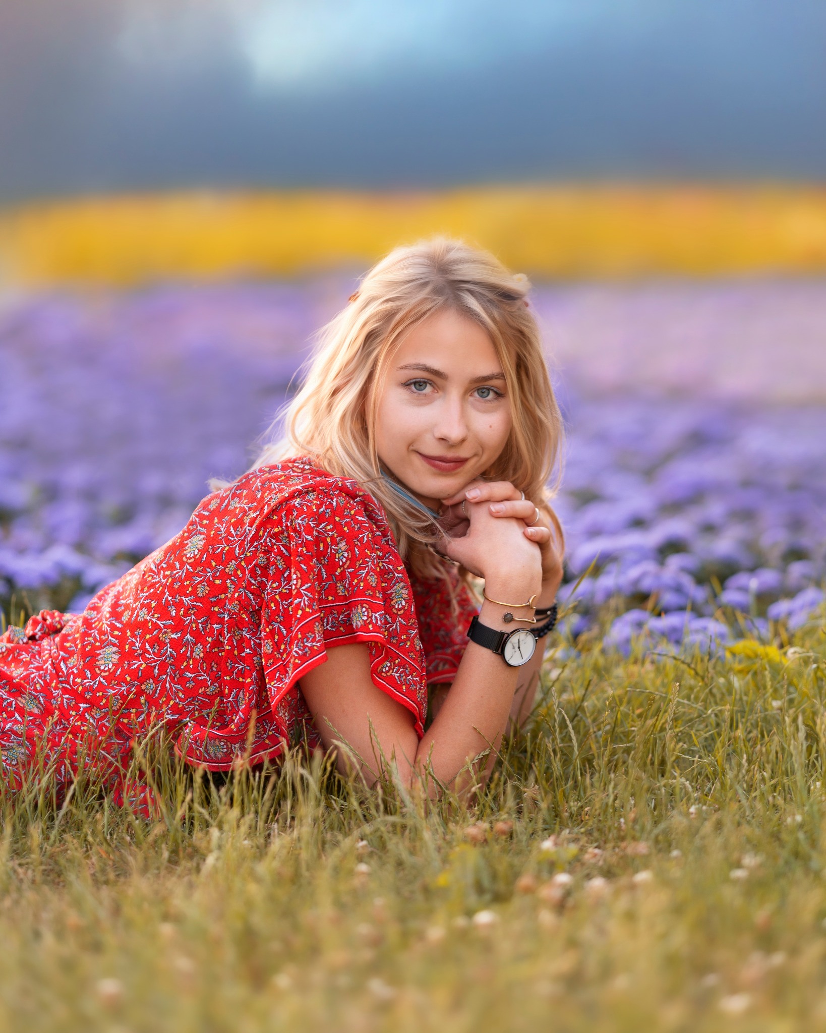 Portret młodej kobiety w czerwonej sukience leżącej na trawie z polem fioletowych kwiatów w tle, z zegarkiem i bransoletkami na nadgarstku.