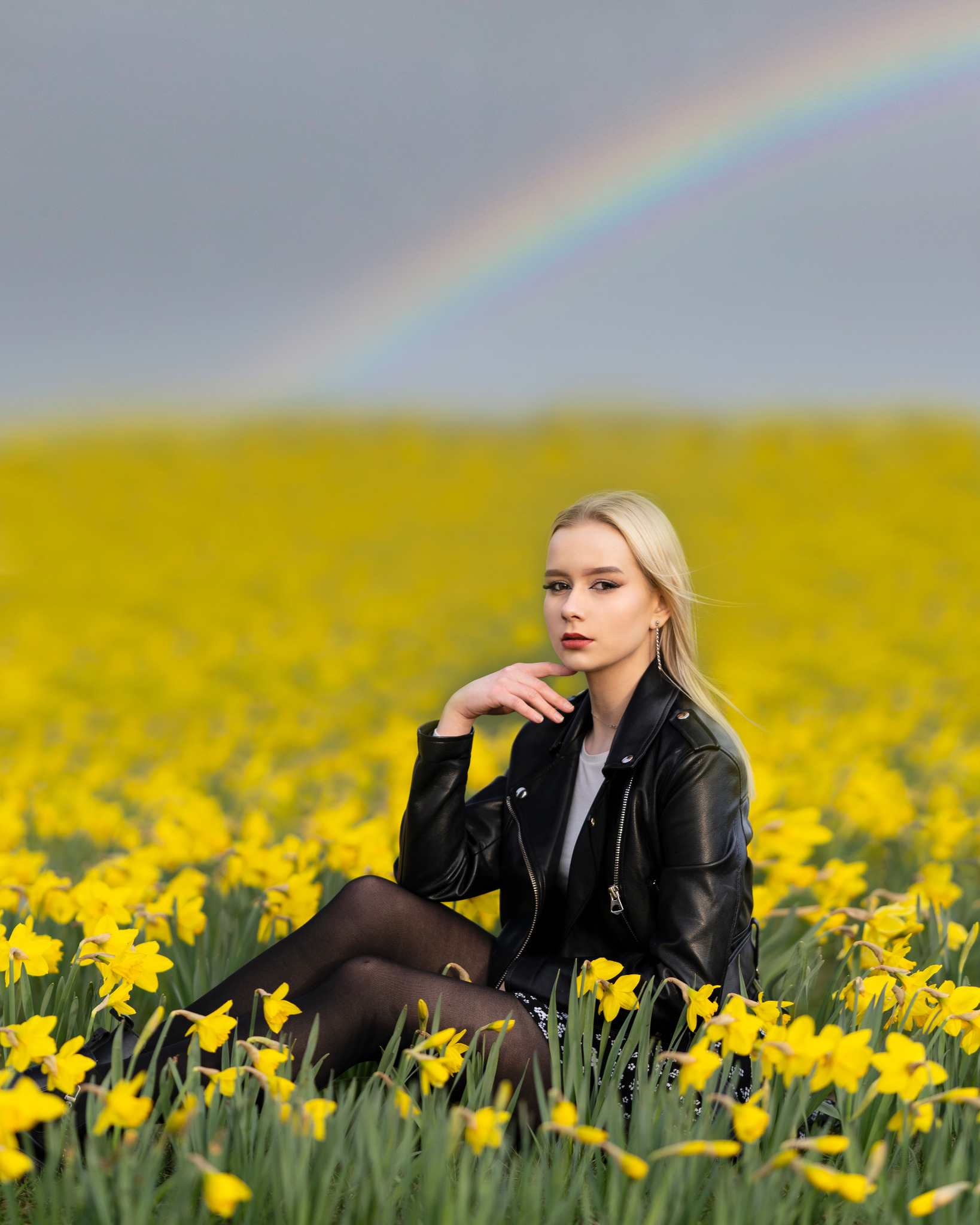 Blondwłosa kobieta w czarnej skórzanej kurtce pozuje siedząc w polu kwitnących żonkili, z tęczą w tle.