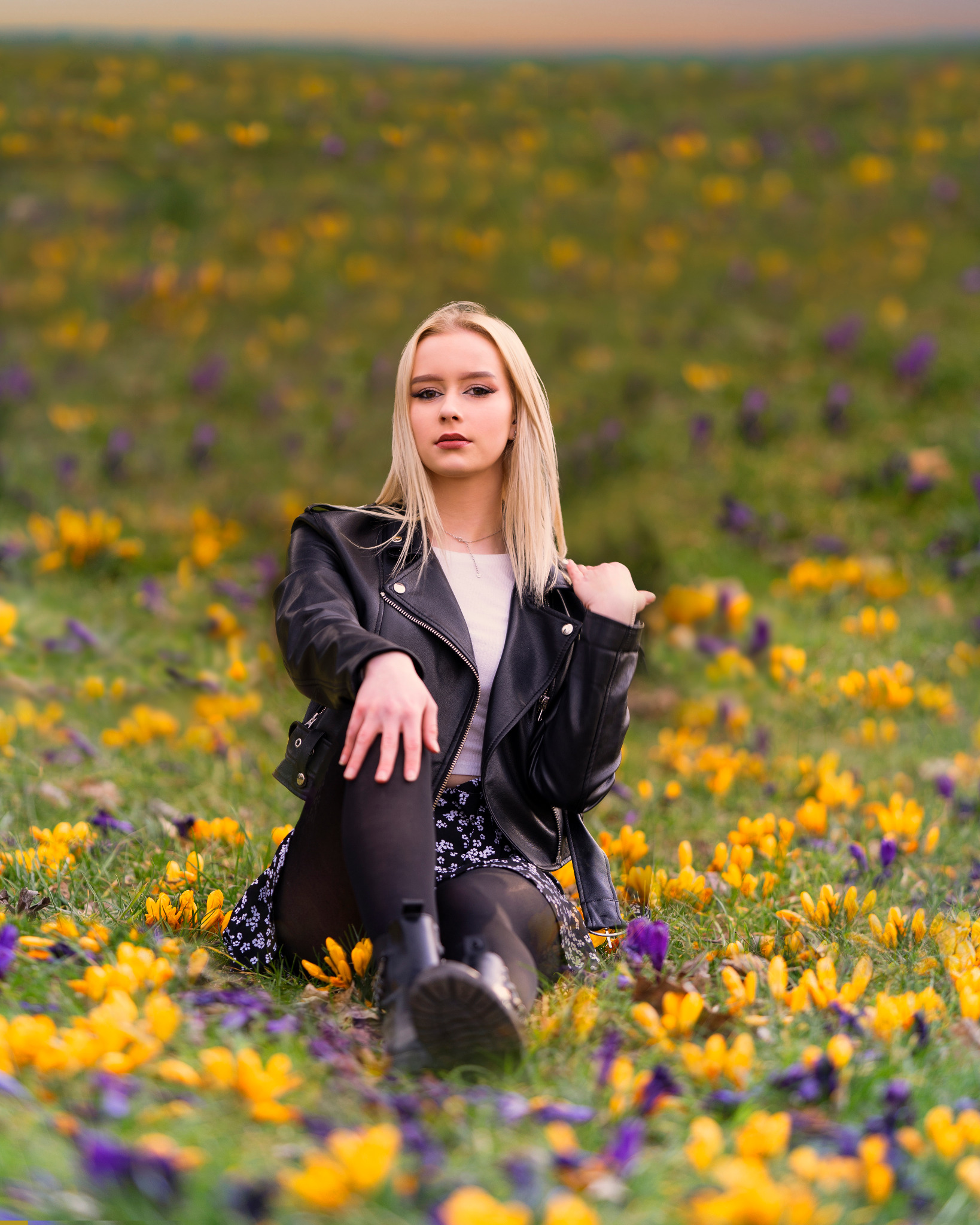 Młoda kobieta w czarnej skórzanej kurtce pozuje siedząc na łące pełnej krokusów w odcieniach żółci i fioletu.