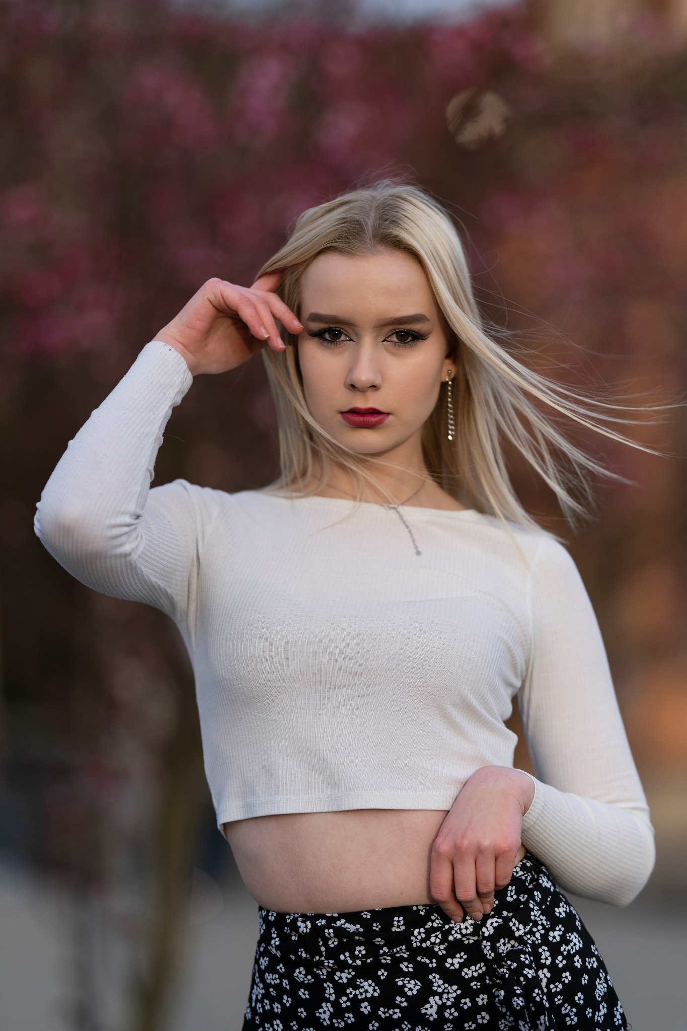 Portret młodej kobiety z blond włosami, w białym topie i kwiecistej spódnicy, pozującej na tle kwitnących drzew.