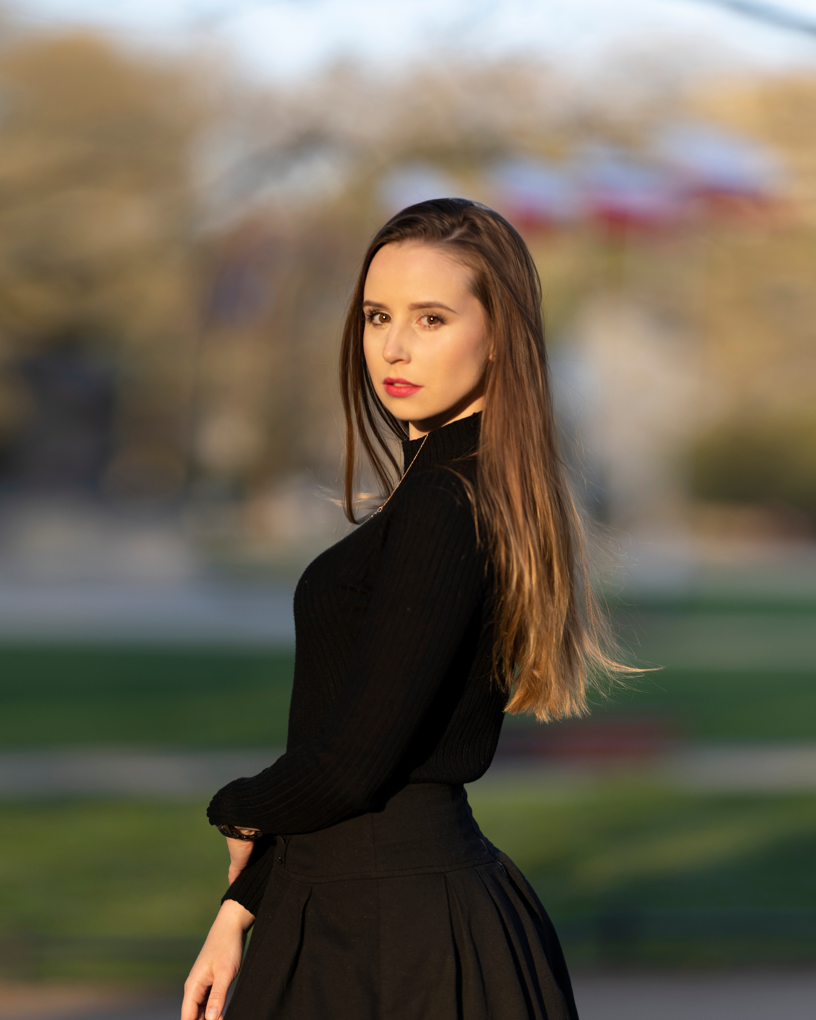 Portret kobiety w czarnej sukience i golfie, z długimi brązowymi włosami, odwróconej przez ramię z delikatnym uśmiechem, na rozmytym tle parkowym w ciepłym świetle.