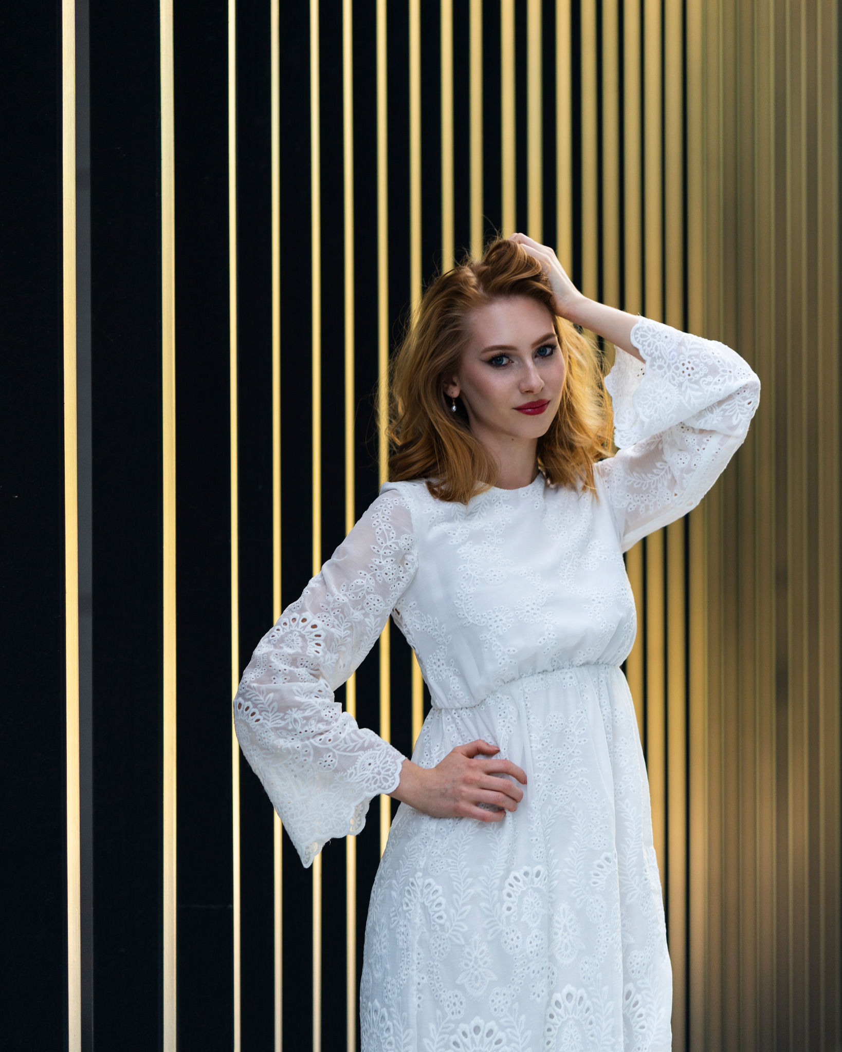 Portret kobiety w białej, koronkowej sukience, pozującej na tle ściany z pionowymi, złotymi pasami.