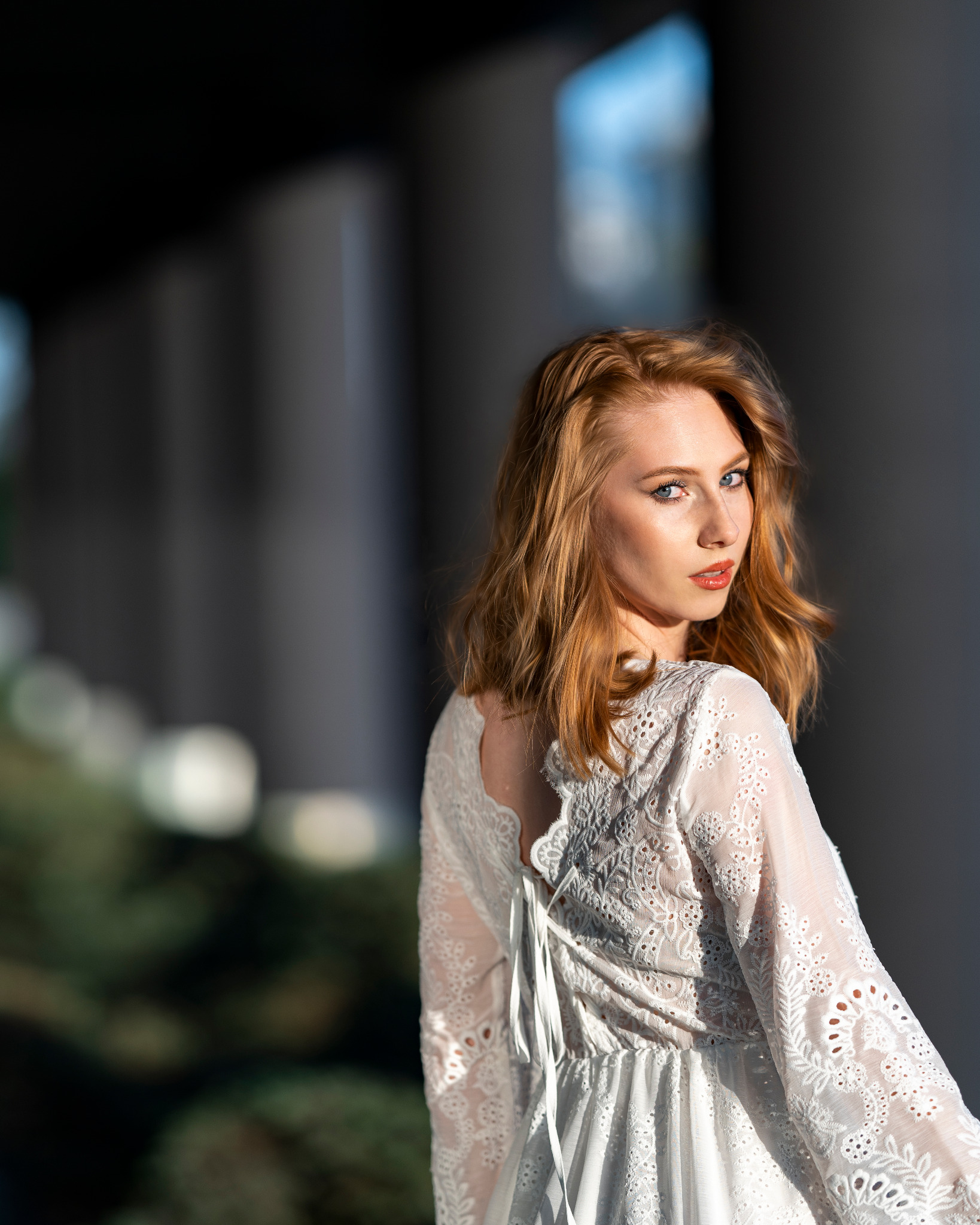 Portret kobiety o rudych włosach w białej koronkowej sukience, odwróconej przez ramię, z głębokim spojrzeniem błękitnych oczu, na tle kolumn w miękkim świetle.