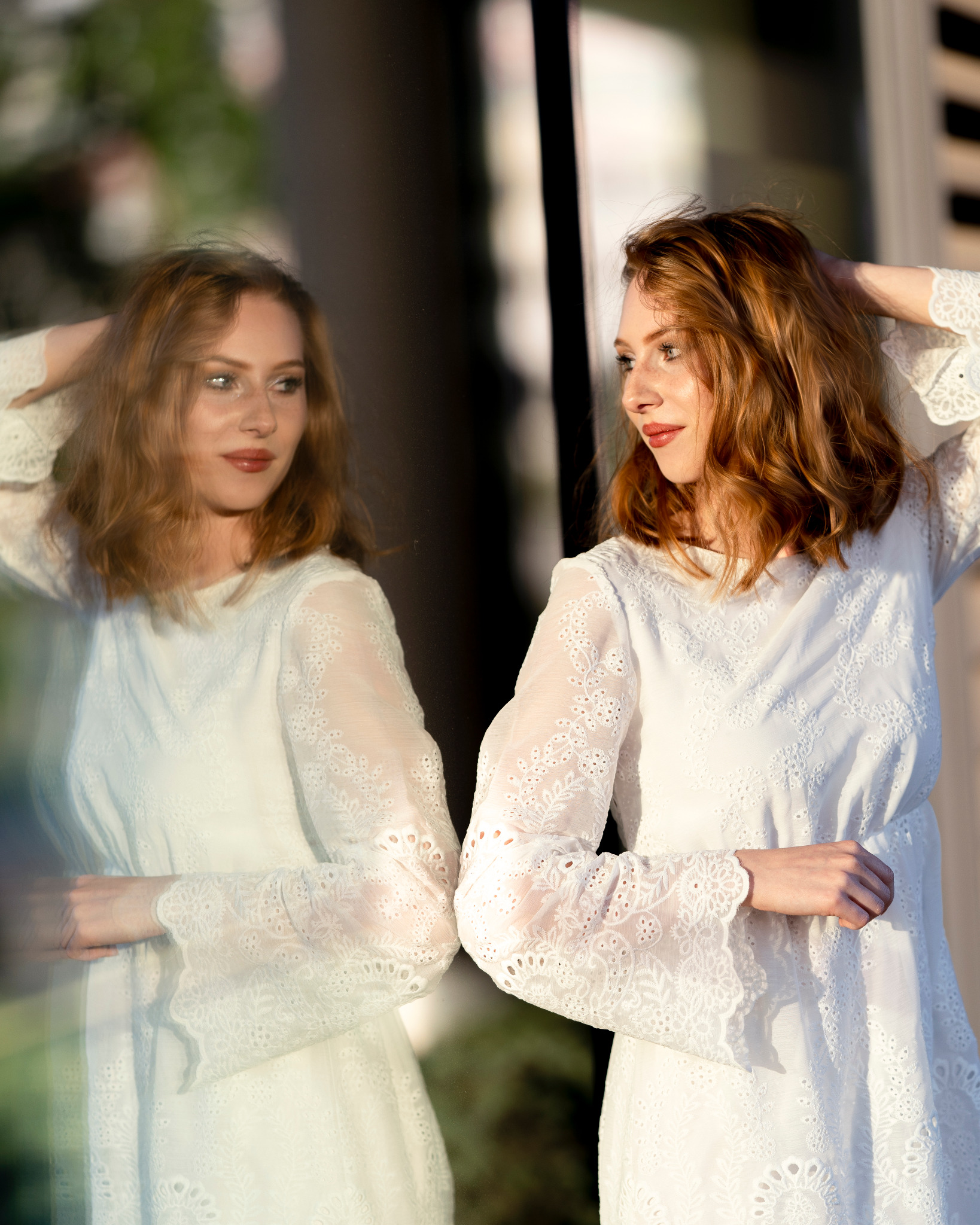 Portret kobiety w białej, koronkowej sukience, odbijającej się w szklanej powierzchni, z delikatnym uśmiechem i rudymi włosami.