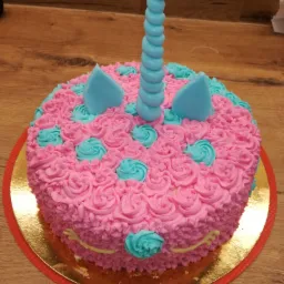 Tort w kształcie jednorożca z różowym kremem w kształcie różyczek i turkusowymi elementami dekoracyjnymi, umieszczony na złotej podkładce na drewnianym blacie.