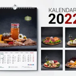 Kalendarz na rok 2022 z ilustracjami produktów spożywczych: miód w słoikach, wędliny, sery i owoce, wiszący na ścianie.