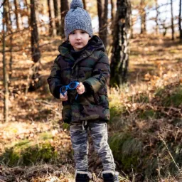 Chłopiec w zimowej czapce i kurtce moro stoi w lesie, trzymając niebieskie okulary.
