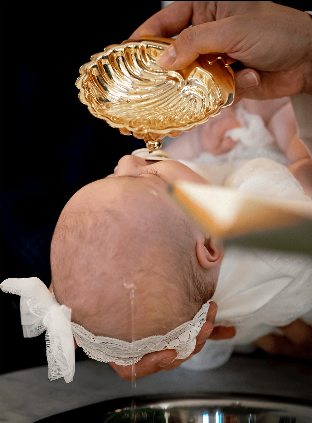 Chrzest dziecka: zbliżenie na głowę dziecka polewaną wodą ze złotej muszli. Delikatne światło podkreśla uroczysty charakter ceremonii.