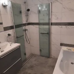 Wykończona łazienka z kabiną prysznicową z dwoma szklanymi ściankami, umywalką z szarą szafką, wanną i płytkami imitującymi marmur z dekoracyjnym pasem mozaiki.