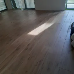 Ułożone panele podłogowe w odcieniach brązu, z widocznym światłem słonecznym padającym na podłogę i częściowo zakrytym oknem w tle.