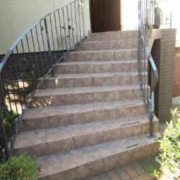 Zewnętrzne schody wejściowe wyłożone brązowymi płytkami ceramicznymi, z metalową, dekoracyjną balustradą z motywem liści, prowadzące do ciemnych drzwi wejściowych.