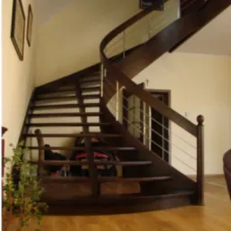 Kręte, drewniane schody z metalowymi poręczami w jasnym wnętrzu, widoczne kwiaty w doniczce i parkiet na podłodze.