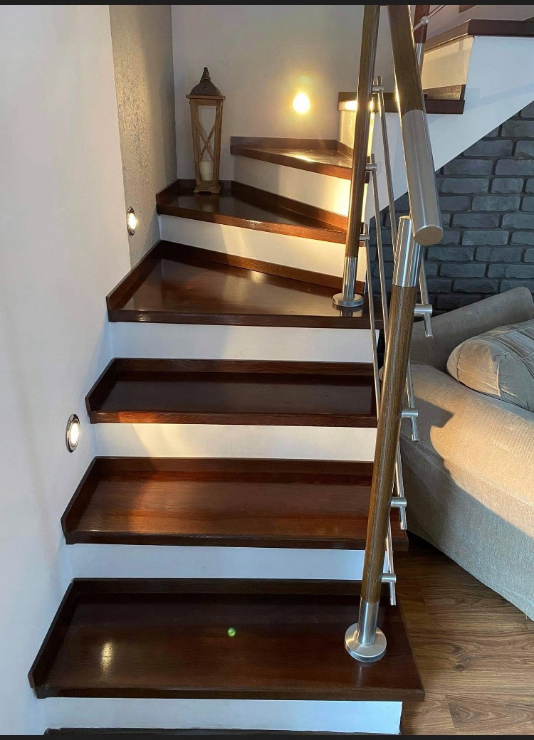 Nowoczesne schody drewniane z podświetleniem LED w ścianie i balustradą ze stali nierdzewnej i drewna, obok szara ściana z cegły i fragment sofy.