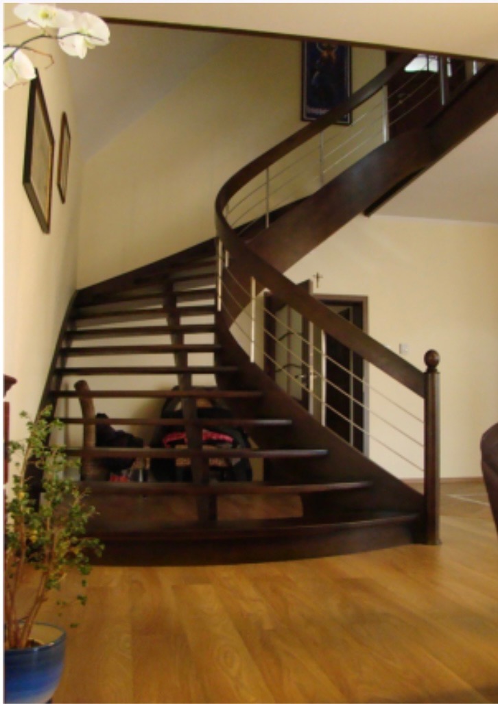 Kręte, drewniane schody z metalowymi poręczami w jasnym wnętrzu, widoczne kwiaty w doniczce i parkiet na podłodze.