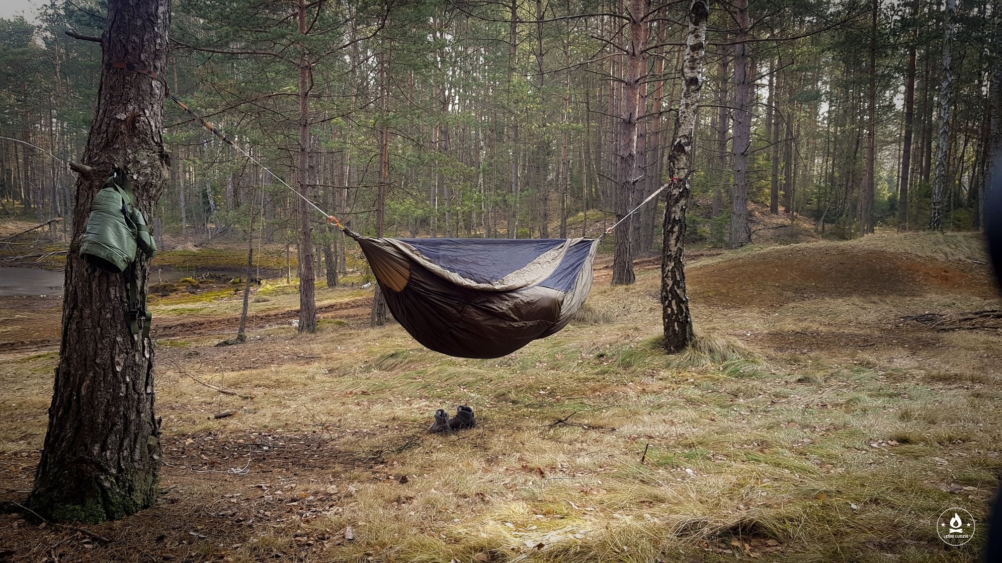 Rozwieszony hamak turystyczny w lesie, z plecakiem zawieszonym na drzewie i parą butów na ziemi.