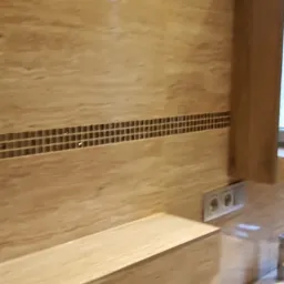 Wykończona łazienka z beżowymi płytkami imitującymi drewno, dekoracyjnym pasem mozaiki, wbudowaną półką i chromowanym przyciskiem spłuczki.