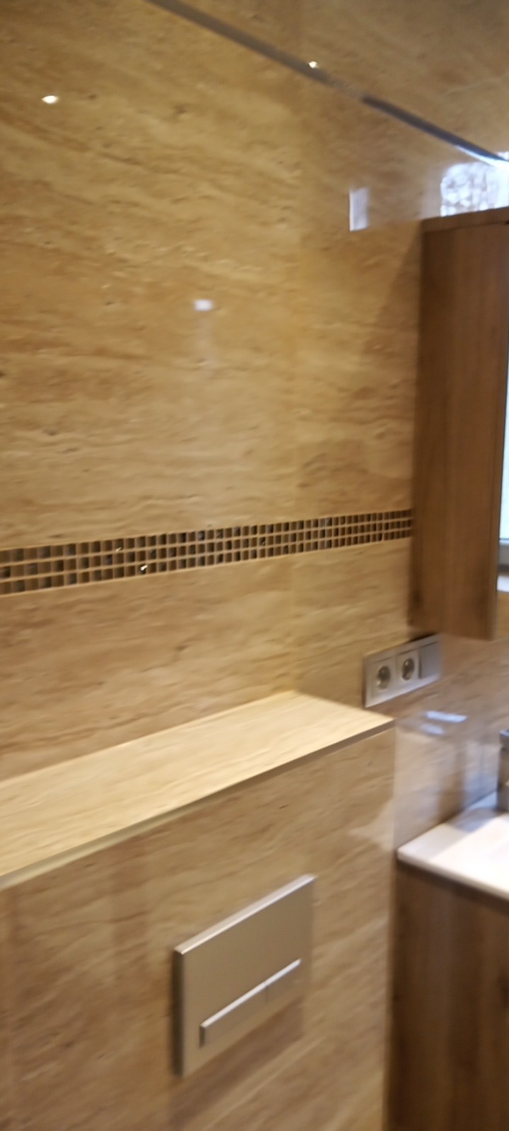 Wykończona łazienka z beżowymi płytkami imitującymi drewno, dekoracyjnym pasem mozaiki, wbudowaną półką i chromowanym przyciskiem spłuczki.