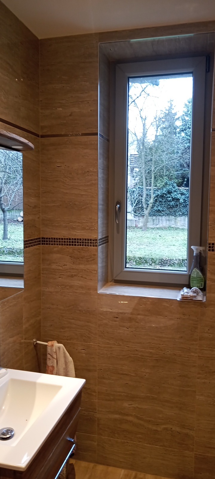 Wykończona łazienka z beżowymi płytkami imitującymi trawertyn, z oknem wychodzącym na ogród, na parapecie płyn do czyszczenia szyb.