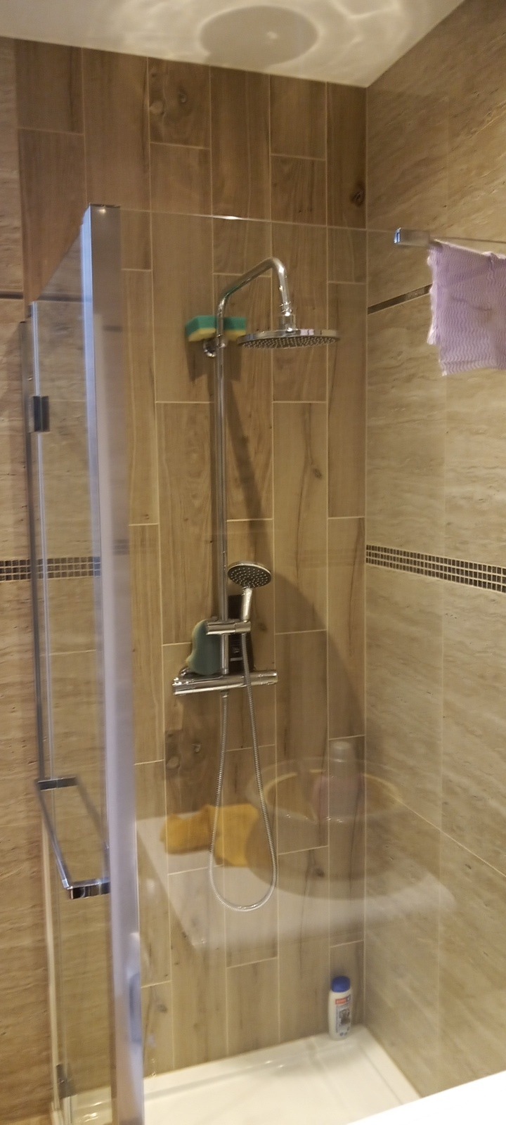 Kabina prysznicowa z deszczownicą i baterią termostatyczną, wykończona płytkami imitującymi drewno, z widocznymi gąbkami do mycia na uchwycie i butelką szamponu w rogu brodzika.