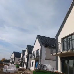 Rząd nowych, bliźniaczych domów jednorodzinnych w trakcie budowy, z zamontowanymi czarnymi balkonami z metalowymi barierkami, widoczne materiały budowlane i kontener na śmieci.