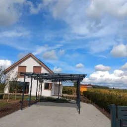 Nowoczesna, metalowa pergola z przezroczystym zadaszeniem na tle domu z brązowymi roletami i błękitnego, zachmurzonego nieba; widoczny fragment ogrodzenia i polna roślinność w tle.