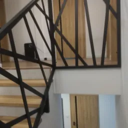 Nowoczesna balustrada metalowa z geometrycznym wzorem na schodach drewnianych, widoczna z dołu, z elementami drzwi w tle.