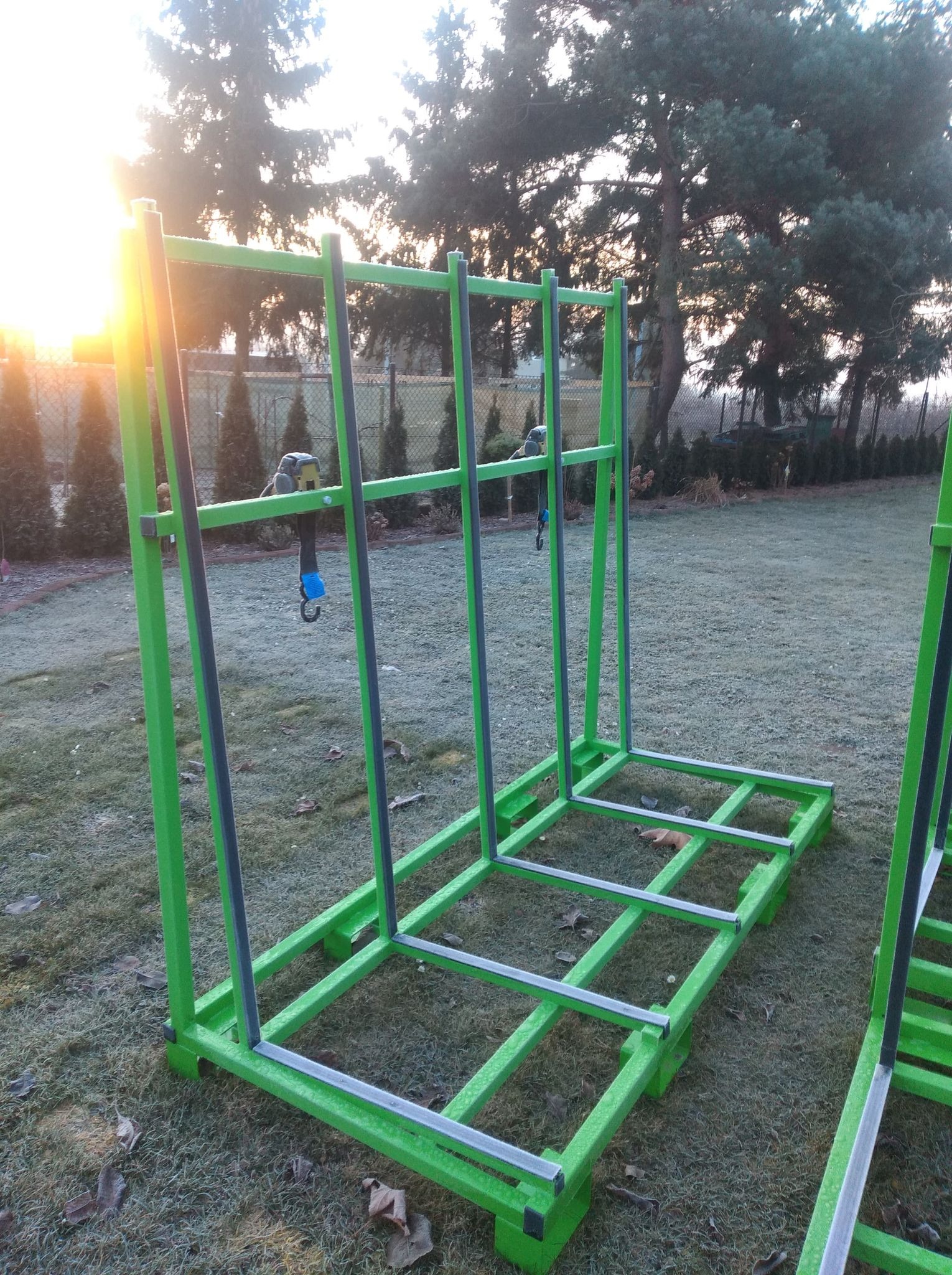 Konstrukcja metalowa w kolorze zielonym i szarym, prawdopodobnie stelaż pod balustradę lub schody, ustawiona na zewnątrz na trawie, w tle drzewa i zachodzące słońce.